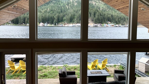 A cabin with large windows offering views of a lake and forest. Inside, there are wicker furniture and a fire pit.