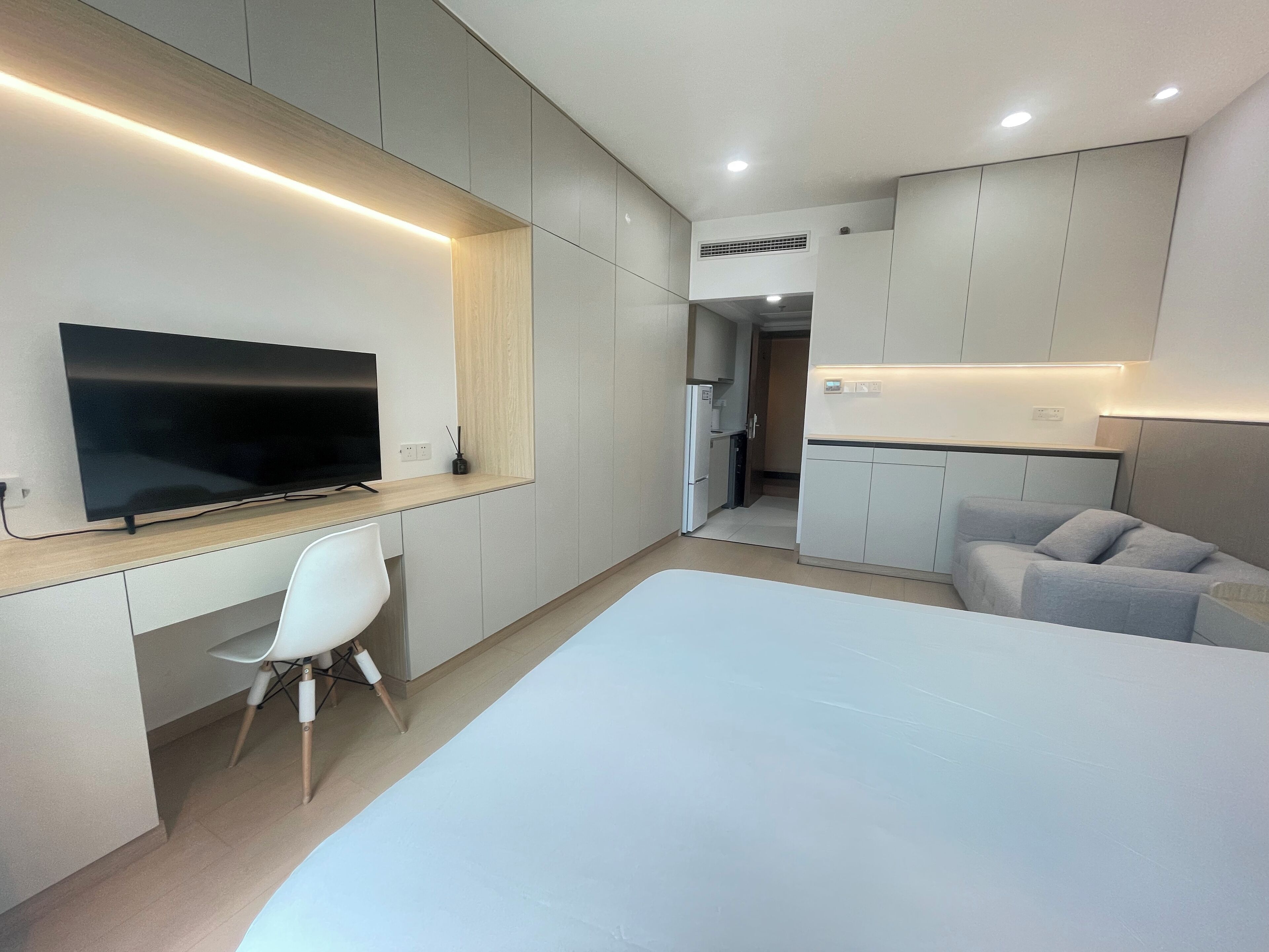 A modern hotel room with a large bed, a desk with a flat-screen TV, a white chair, and a sofa.