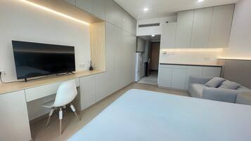 A modern hotel room with a large bed, a desk with a flat-screen TV, a white chair, and a sofa.