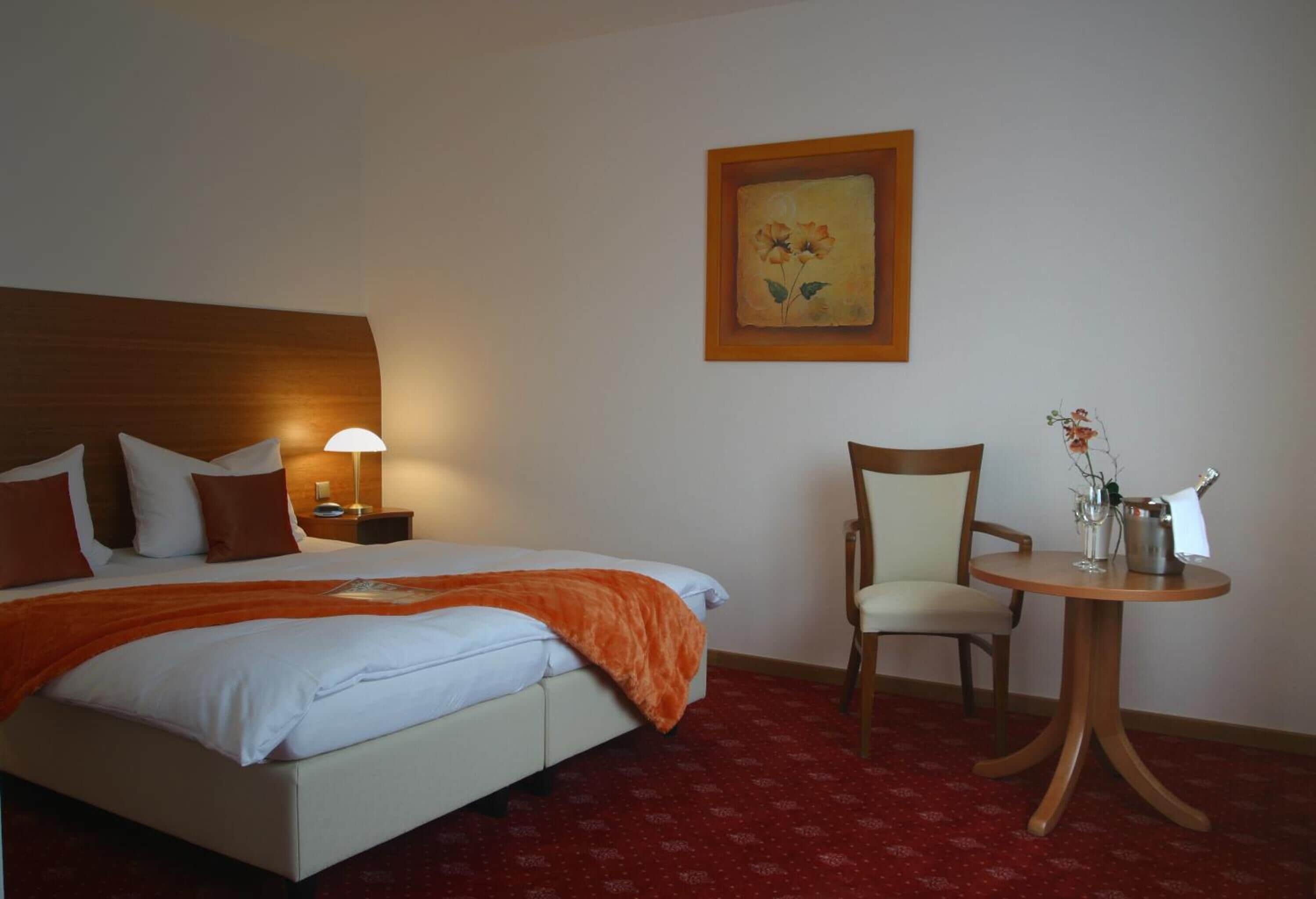 Quarto de hotel com cama, mesas de cabeceira, uma cadeira, uma pequena mesa com um vaso e um quadro na parede.