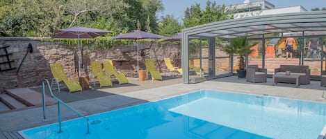 Ein Swimmingpool mit Liegestühlen und Sonnenschirmen, umgeben von einer Steinmauer und Grünpflanzen.