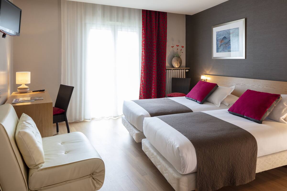 Ein Hotelzimmer mit zwei Betten, einem Schreibtisch, einem Sessel, einem Fernseher und einem Fenster mit Vorhängen.