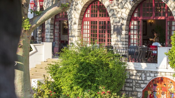 A restaurant with stone facade, red-framed windows, outdoor seating, and a balcony. - Hotel La Chaumiere (Quillan)
