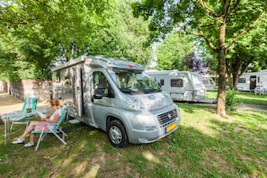 A silver camper van parked in a grassy area with a woman sitting on a chair nearby. - Camping Vicenza (Vicenza)