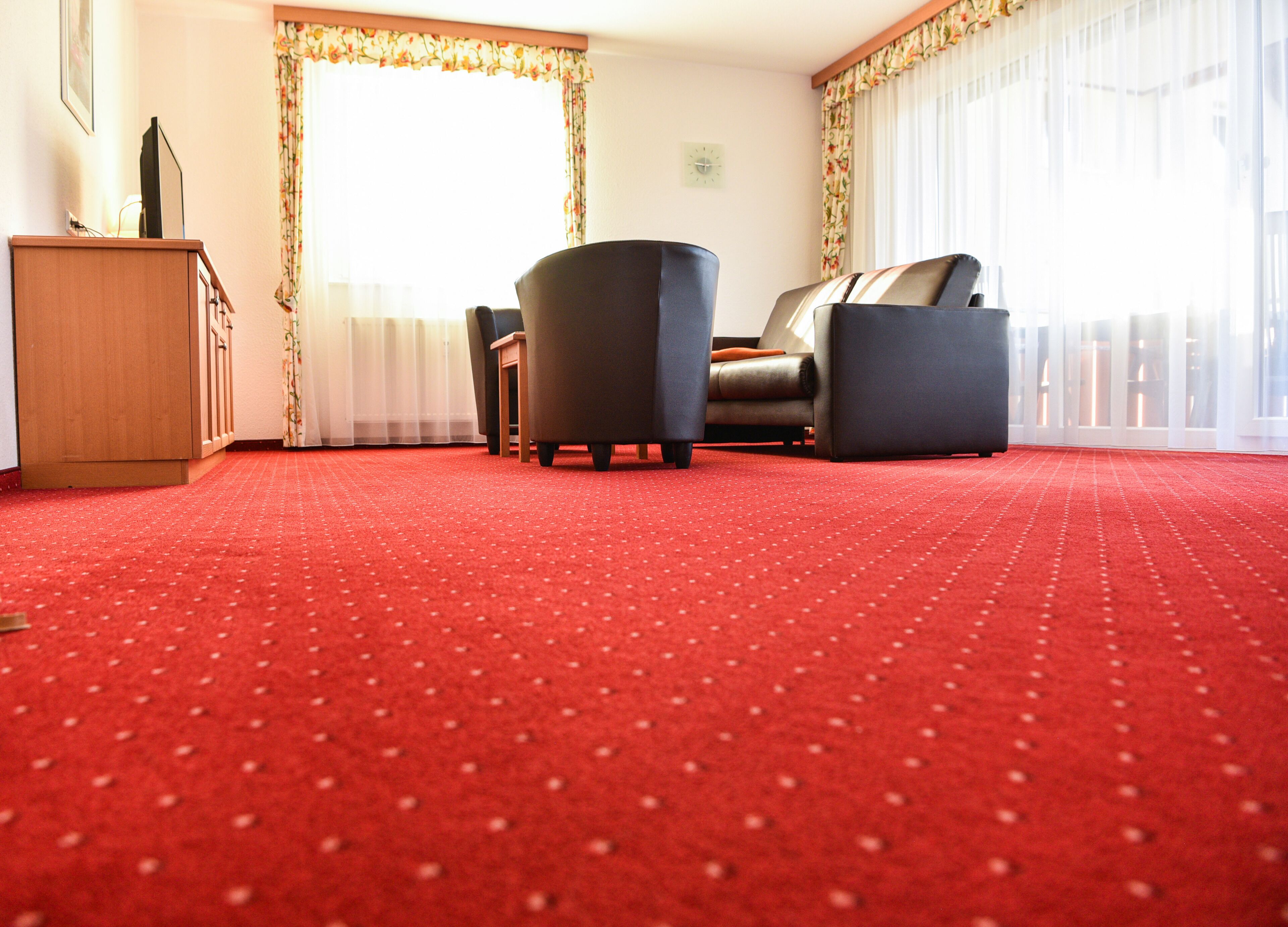 Une pièce avec un tapis rouge, un canapé, un fauteuil, un meuble en bois et une fenêtre avec des rideaux.