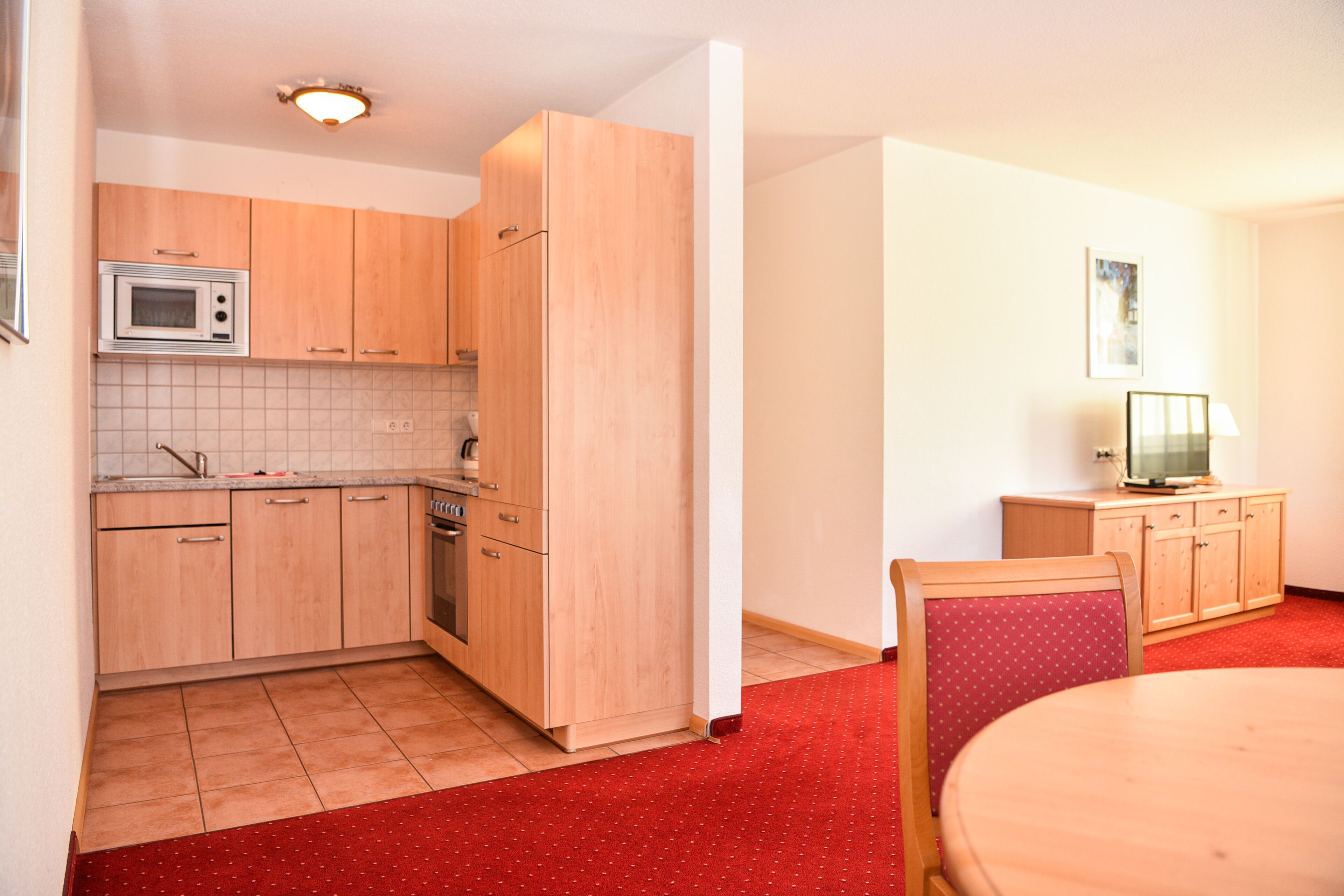 A compact kitchen with wooden cabinets, a microwave, and a red carpeted floor.