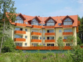 Un hotel de varios pisos con balcones, una cerca verde y abundante vegetación alrededor.