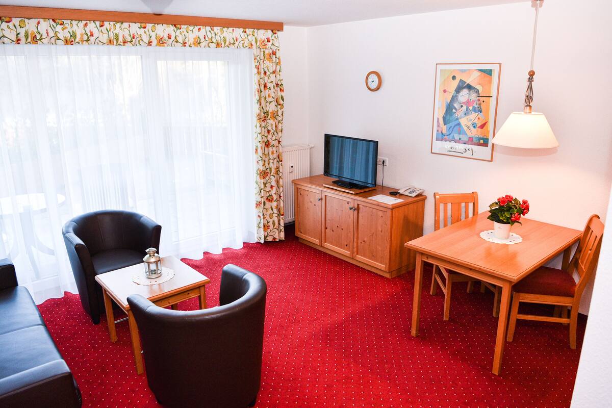 Una habitación con alfombra roja, una mesa de comedor de madera con sillas, un televisor sobre un mueble de madera, un sofá y una ventana con cortinas.