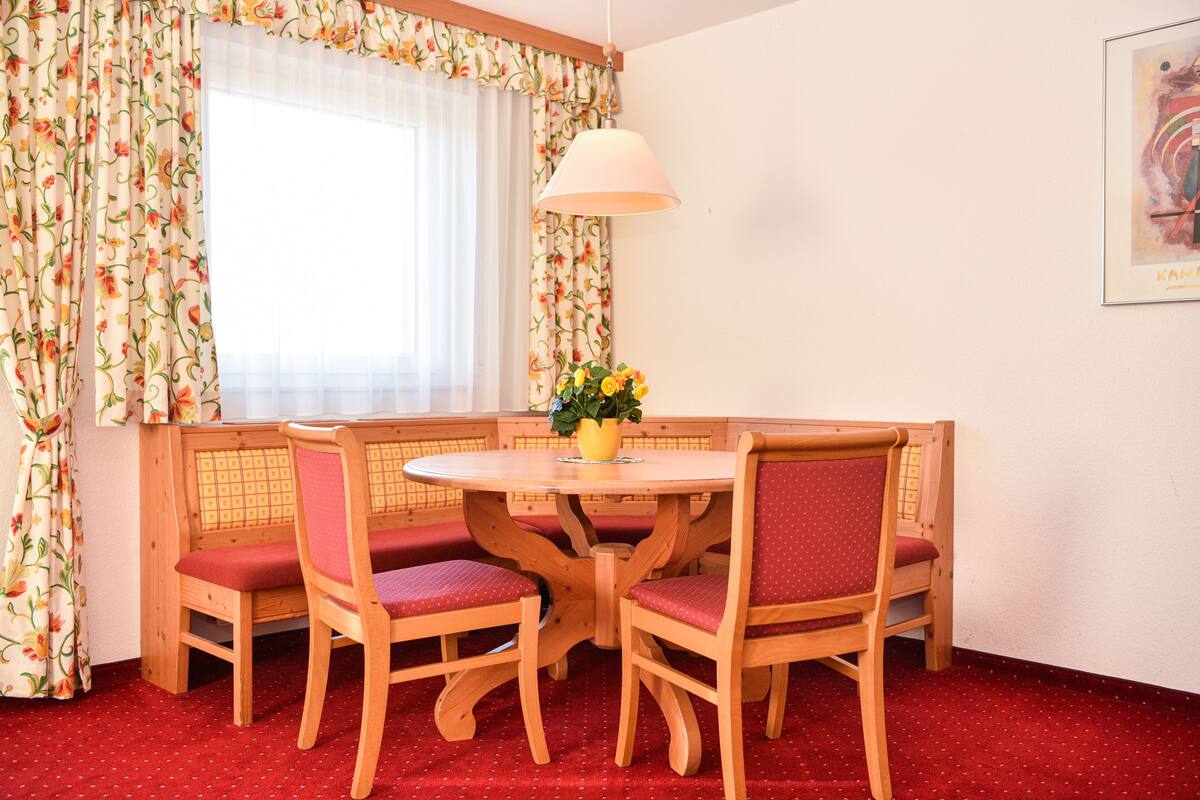 Un comedor con una mesa redonda de madera, cuatro sillas con cojines rojos, un jarrón con flores y una lámpara colgante.