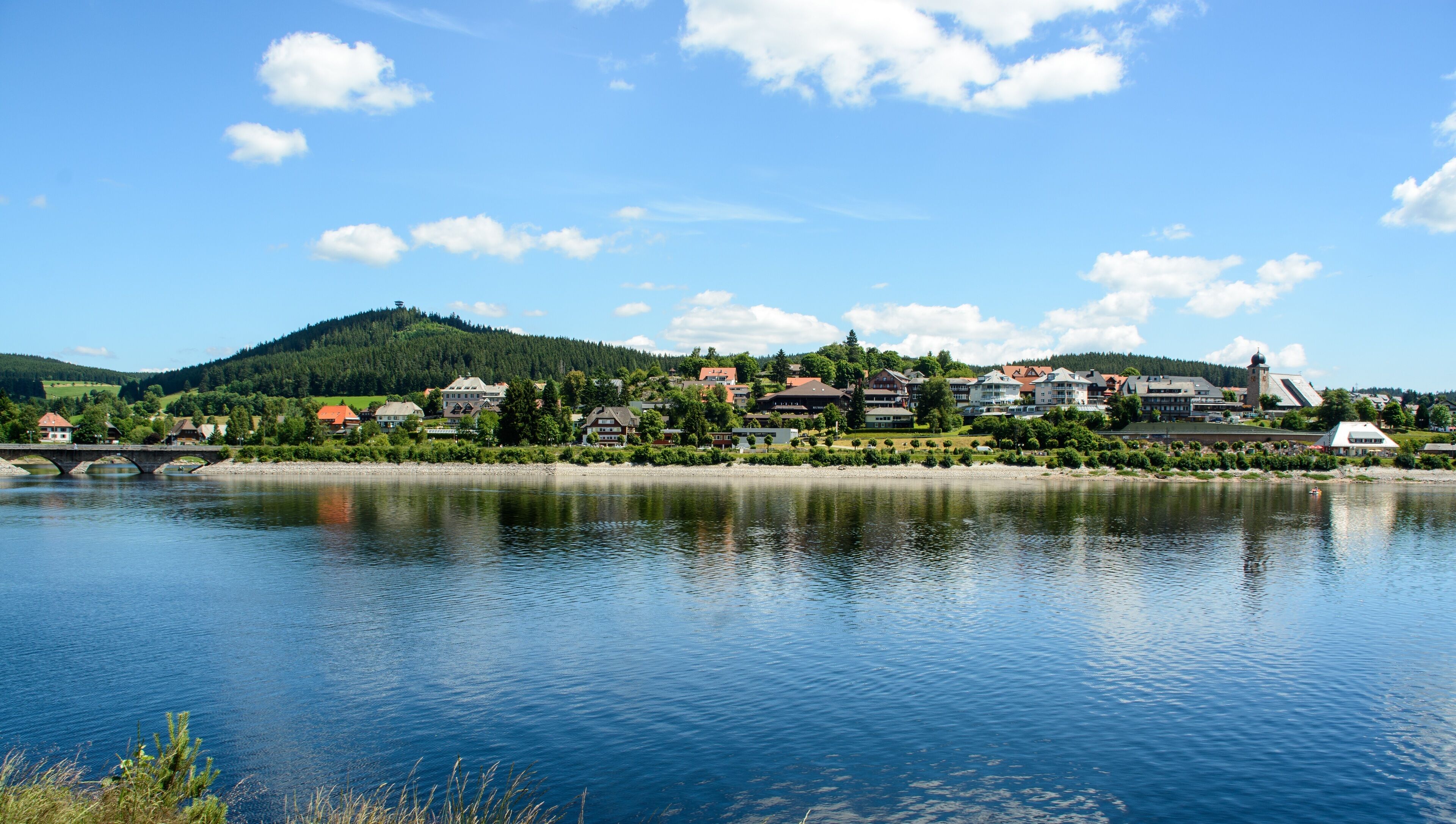 A lakeside town with a bridge, buildings, and a hill in the background.