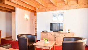 A room with wooden ceiling beams, a red carpet, a wooden table with a vase, two leather chairs, a television on a wooden cabinet, and a framed picture on the wall. - Aparthotel Sonnenburg (Schluchsee)