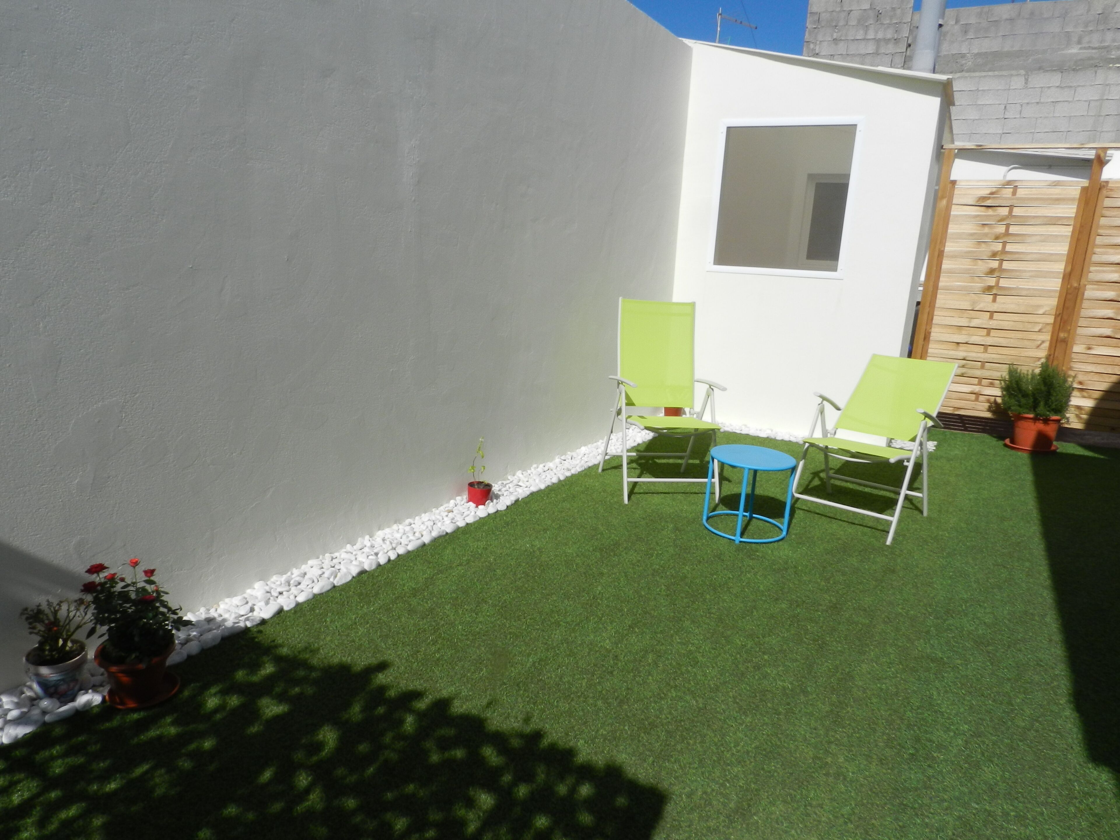 Un petit espace patio avec du gazon synthétique, deux chaises vertes, une table bleue et des plantes en pot.