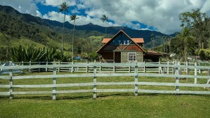 A wooden cabin with a red roof, surrounded by a white fence, set against a backdrop of lush green hills and a mountain range under a partly cloudy sky.