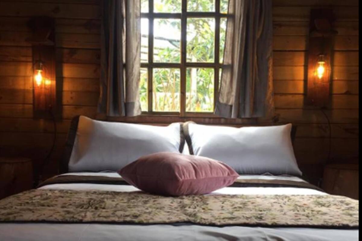 Una habitación de cabaña de madera con una cama, dos apliques de pared y una ventana con cortinas.
