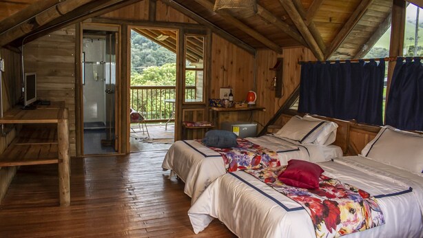 A wooden cabin with two beds, a desk, and a balcony with a view of the forest.