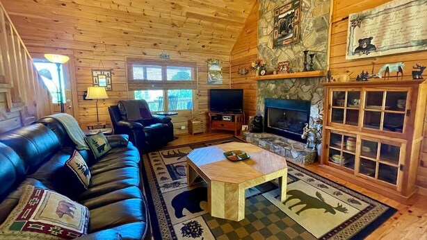 A cozy cabin interior with a wooden ceiling, a stone fireplace, a leather sofa, a television, and a checkered rug. - Sunrise Sensation - Great Views, Hot Tub, Gameroom, Close to Town, WIFI, Covered Porch (West Jefferson)