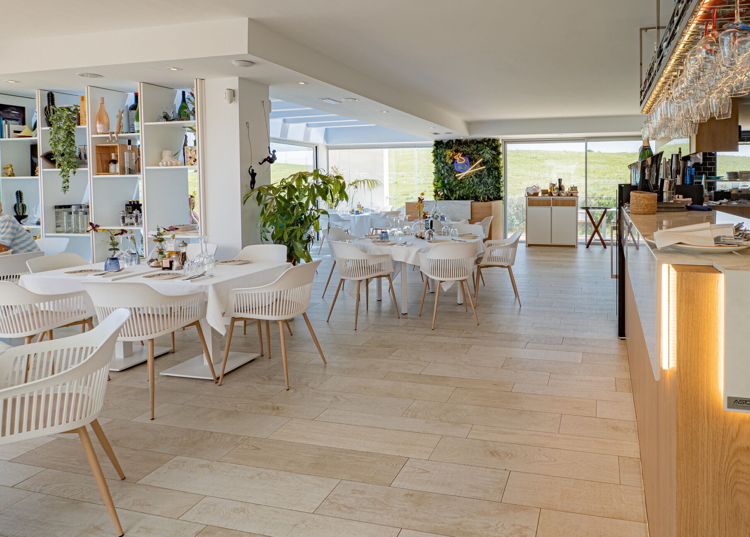 A spacious dining area with white tables and chairs, a large plant, and a view of the outdoors.