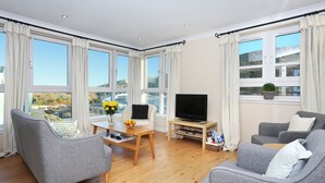 A modern living room with a flat-screen TV, grey sofas, wooden flooring, and large windows offering a view of the outdoors. - Edinburgh Arthur Seat View Apartment (Edinburgh)