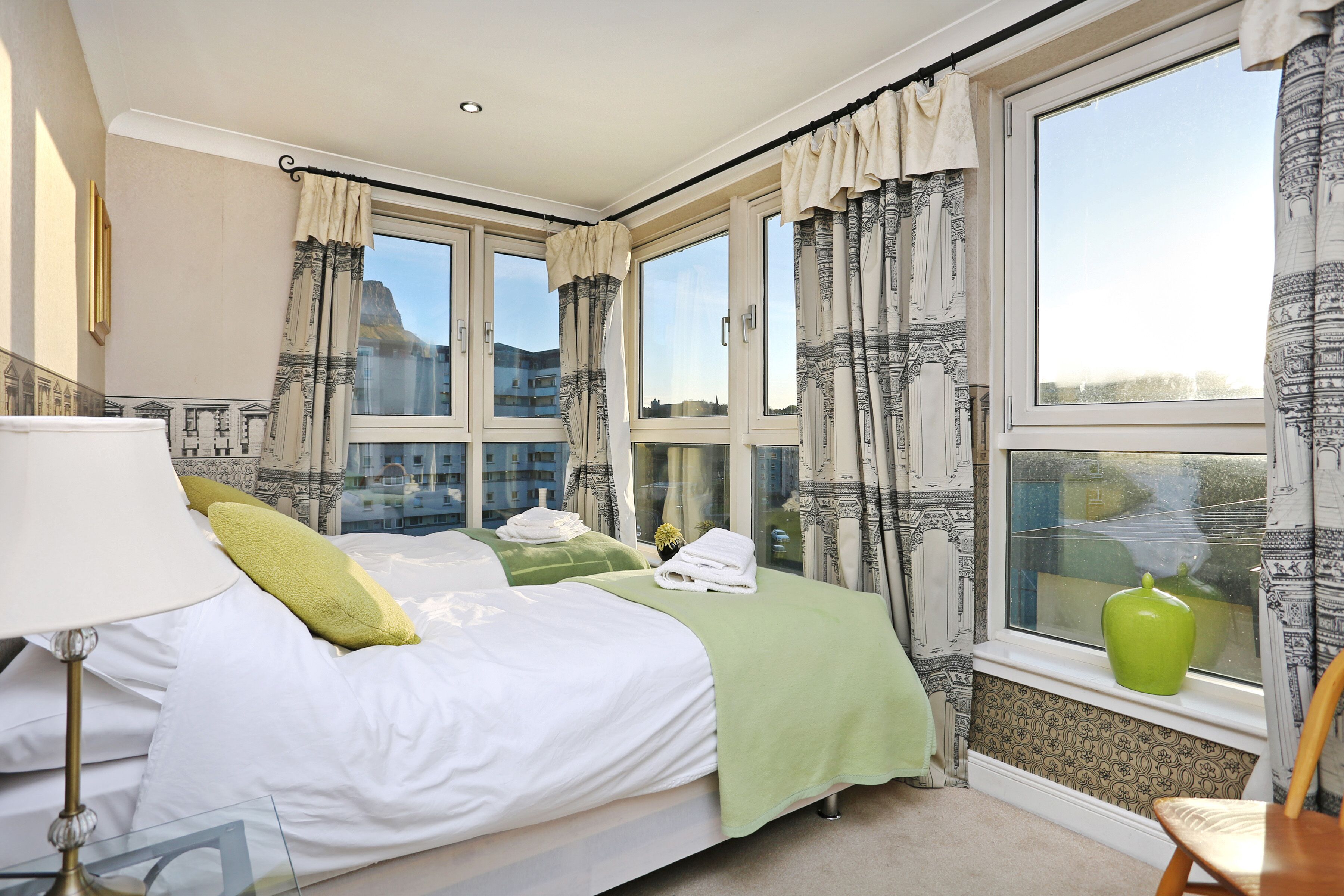 Une chambre d’hôtel avec deux lits, une grande fenêtre avec des rideaux, un vase vert posé sur le rebord de la fenêtre et une chaise.