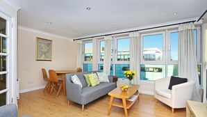 A modern living room with a grey sofa, a white armchair, and a wooden coffee table. There are large windows with curtains, a dining table with chairs, and a vase with flowers on the table. - Edinburgh Arthur Seat View Apartment (Edinburgh)