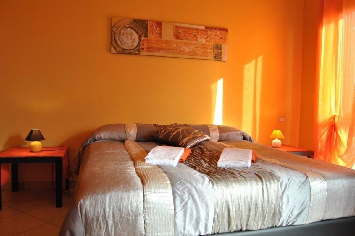 Un dormitorio con una cama grande, dos mesitas de noche con lámparas, un cuadro en la pared y cortinas color naranja.