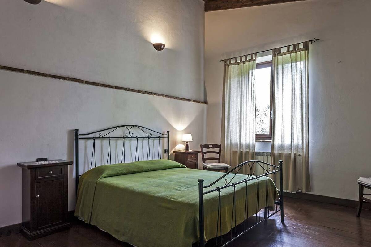 A single bed with a metal frame and green bedding, a bedside table with a lamp, a chair, and a window with curtains.
