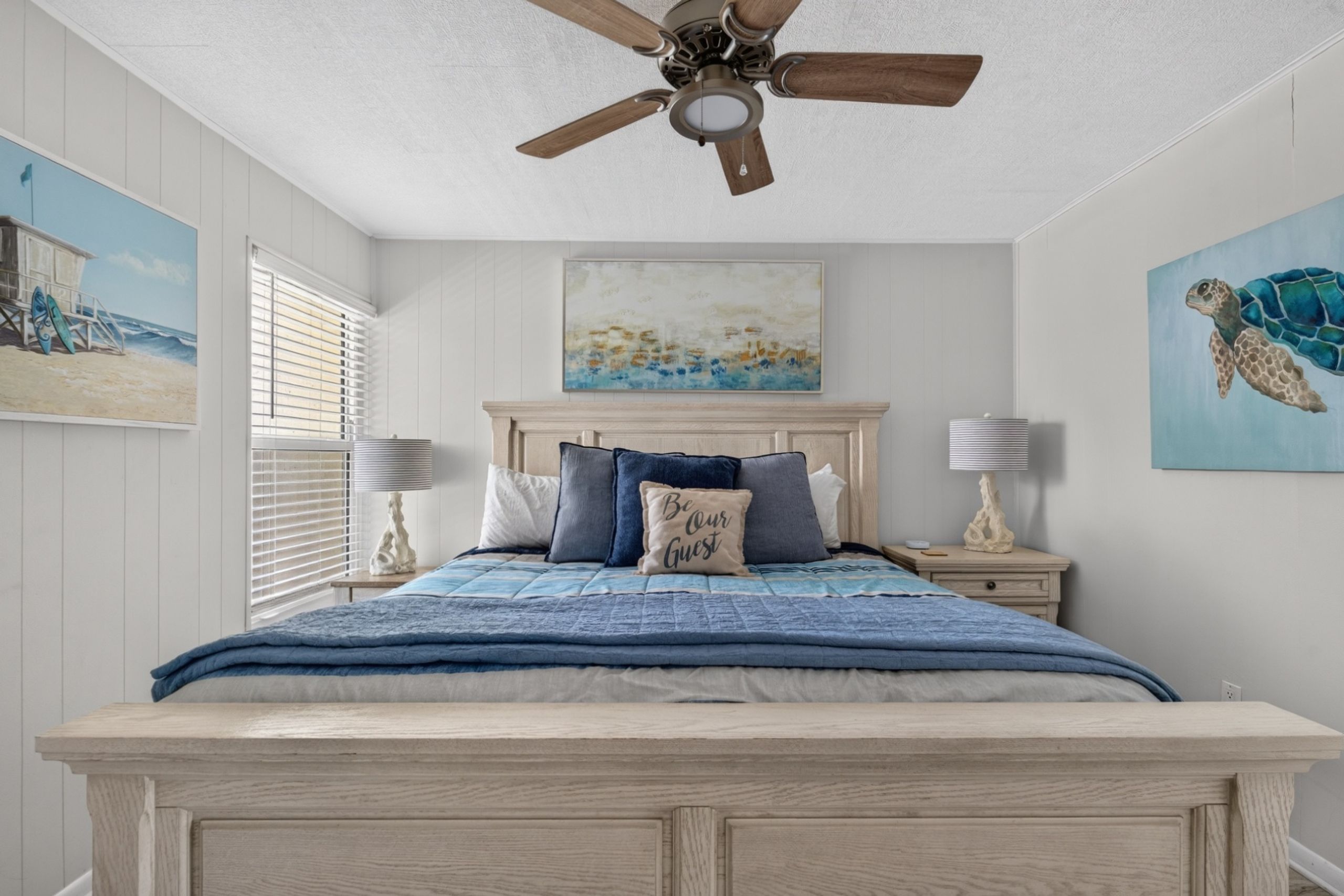 A bedroom with a bed, a ceiling fan, a painting of a beach scene, and a painting of a sea turtle.