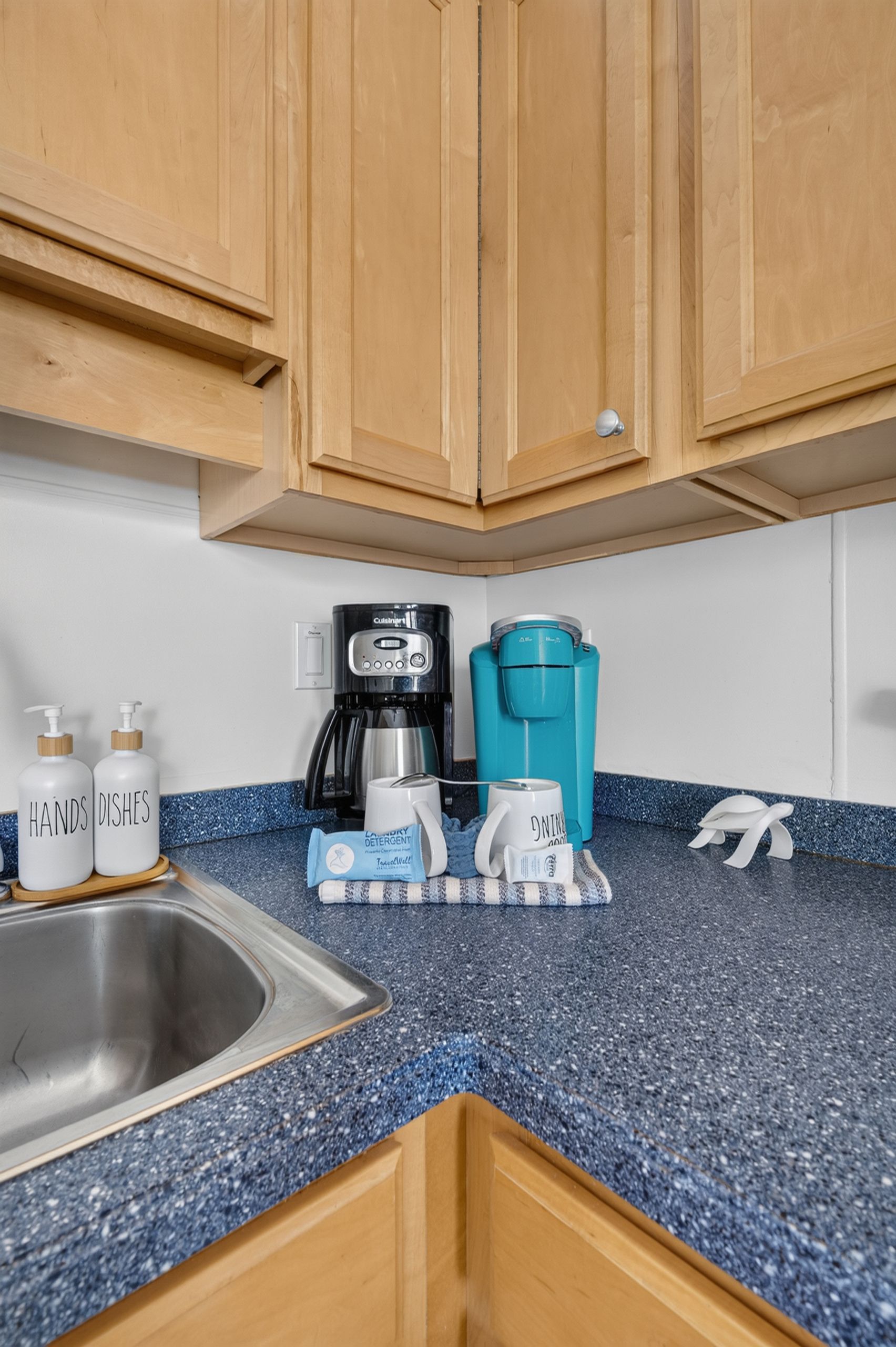 Een keuken met houten kastjes, een roestvrijstalen spoelbak, een koffiezetapparaat, een waterkoker en een mok.