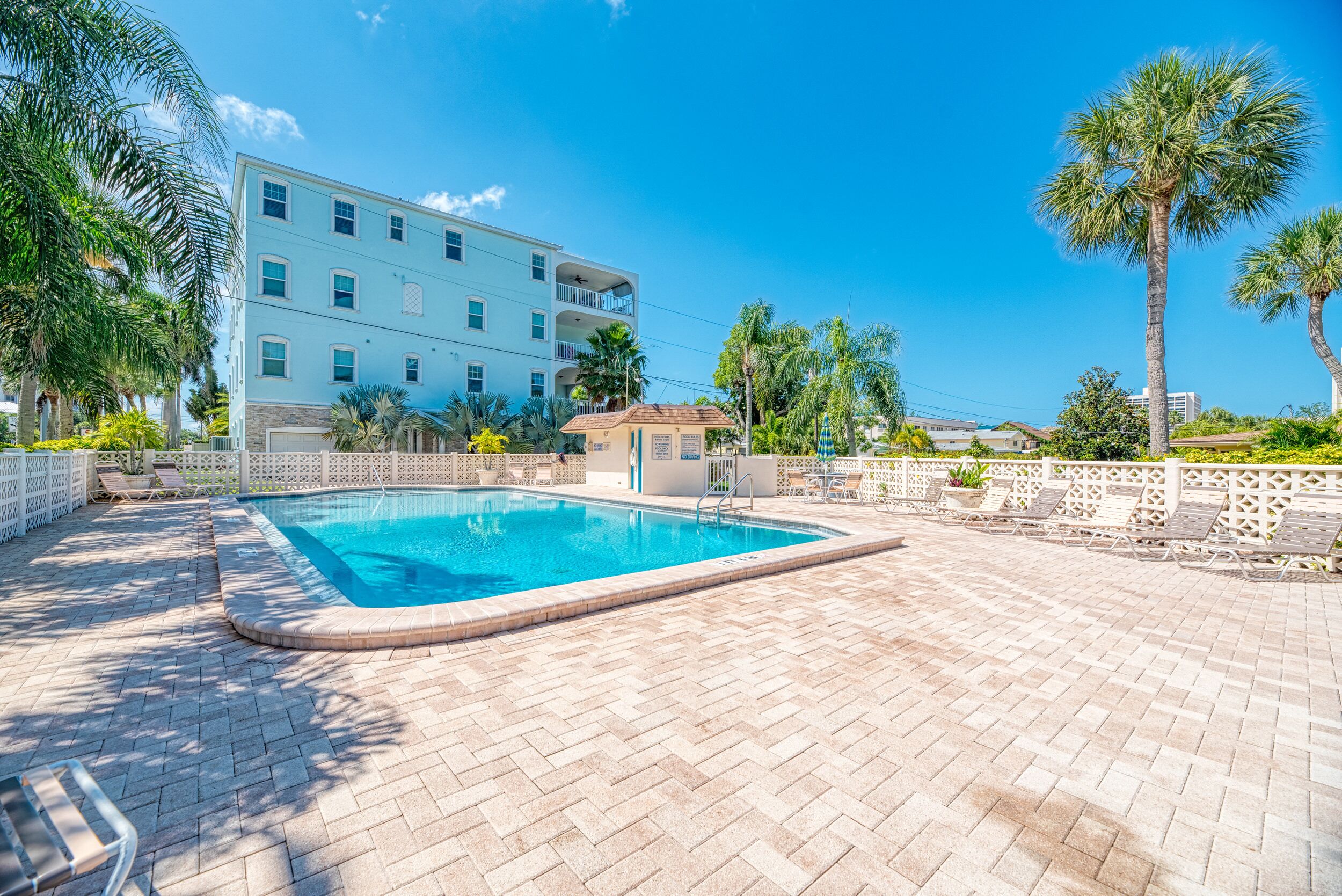 A swimming pool with a tiled deck, surrounded by palm trees and a building with balconies.