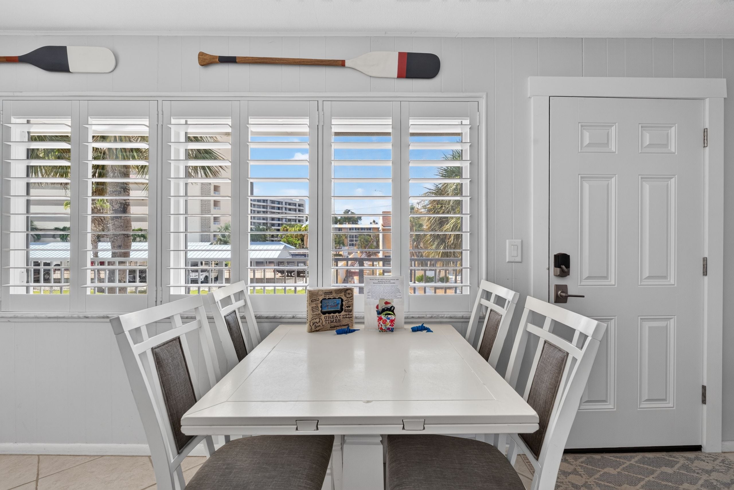 A dining area with a white table and chairs, a window with white shutters, and a door with a handle.