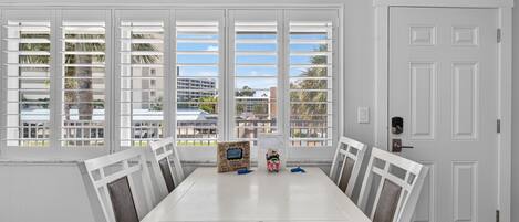 A dining area with a white table and chairs, a window with white shutters, and a door with a handle.