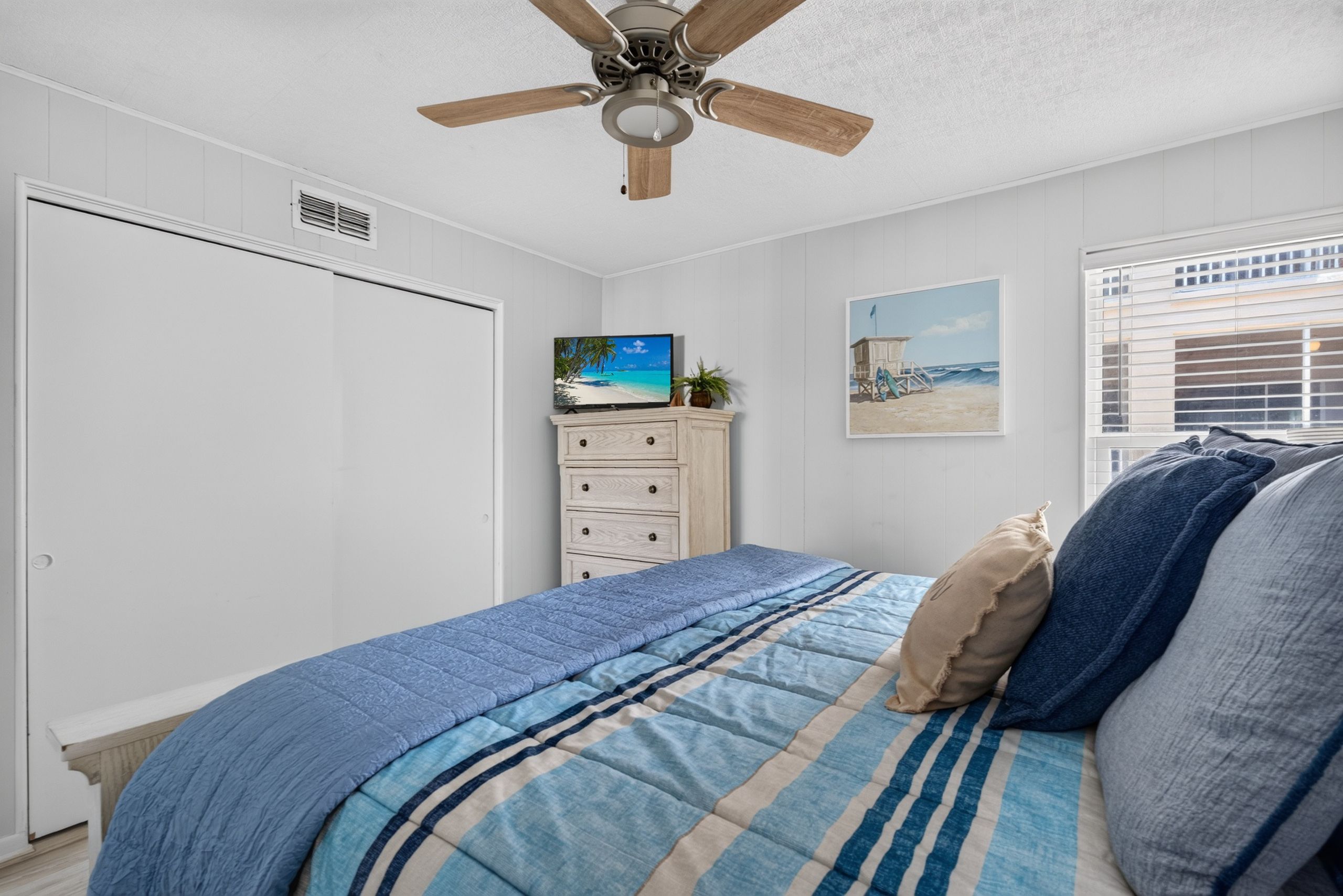 A bedroom with a bed, a ceiling fan, a dresser, a television, and a window with blinds.