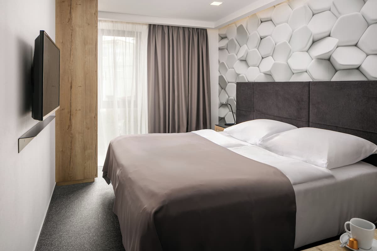 A hotel room with a bed, a flat-screen TV mounted on the wall, a wooden door, and a window with curtains.