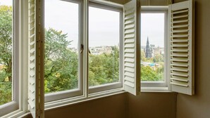 Quarto com três janelas, cada uma com persianas brancas, com vista para uma paisagem urbana com áreas verdes e edifícios.