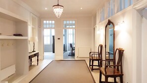 Um corredor com um candelabro, cadeiras de madeira, um espelho e vista para outro quarto.