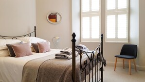 Uma cama bem arrumada com almofadas, um abajur e uma cadeira em um quarto com uma janela grande e um espelho na parede.