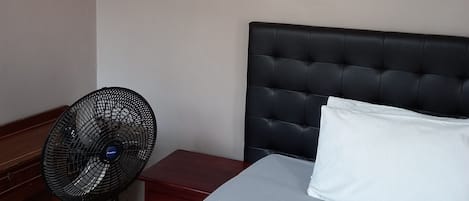 A hotel room with a bed, a nightstand, a desk, and a wall-mounted fan.