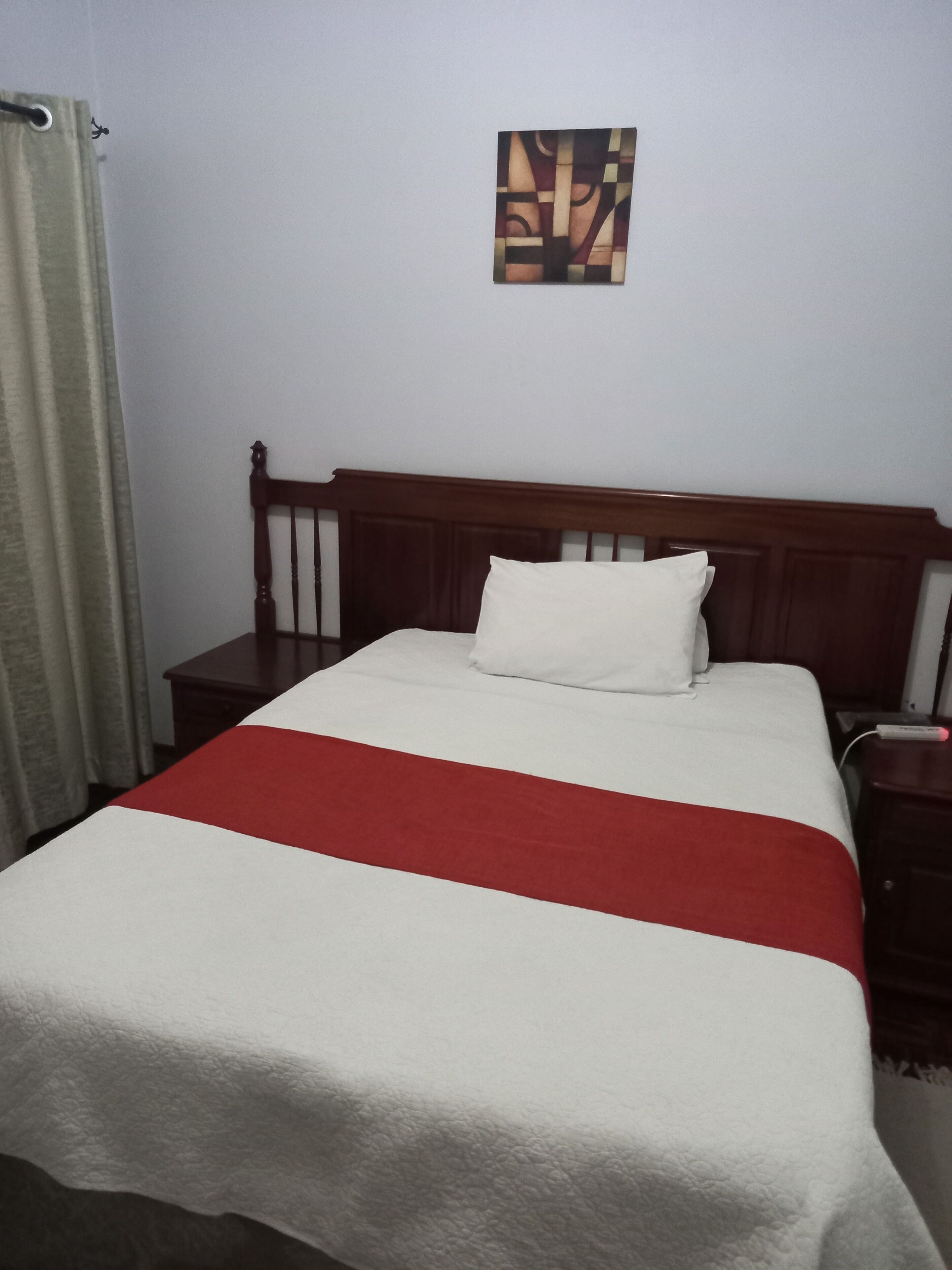 Una cama bien hecha con una colcha blanca y roja, un cabecero de madera y un pequeño cuadro en la pared.