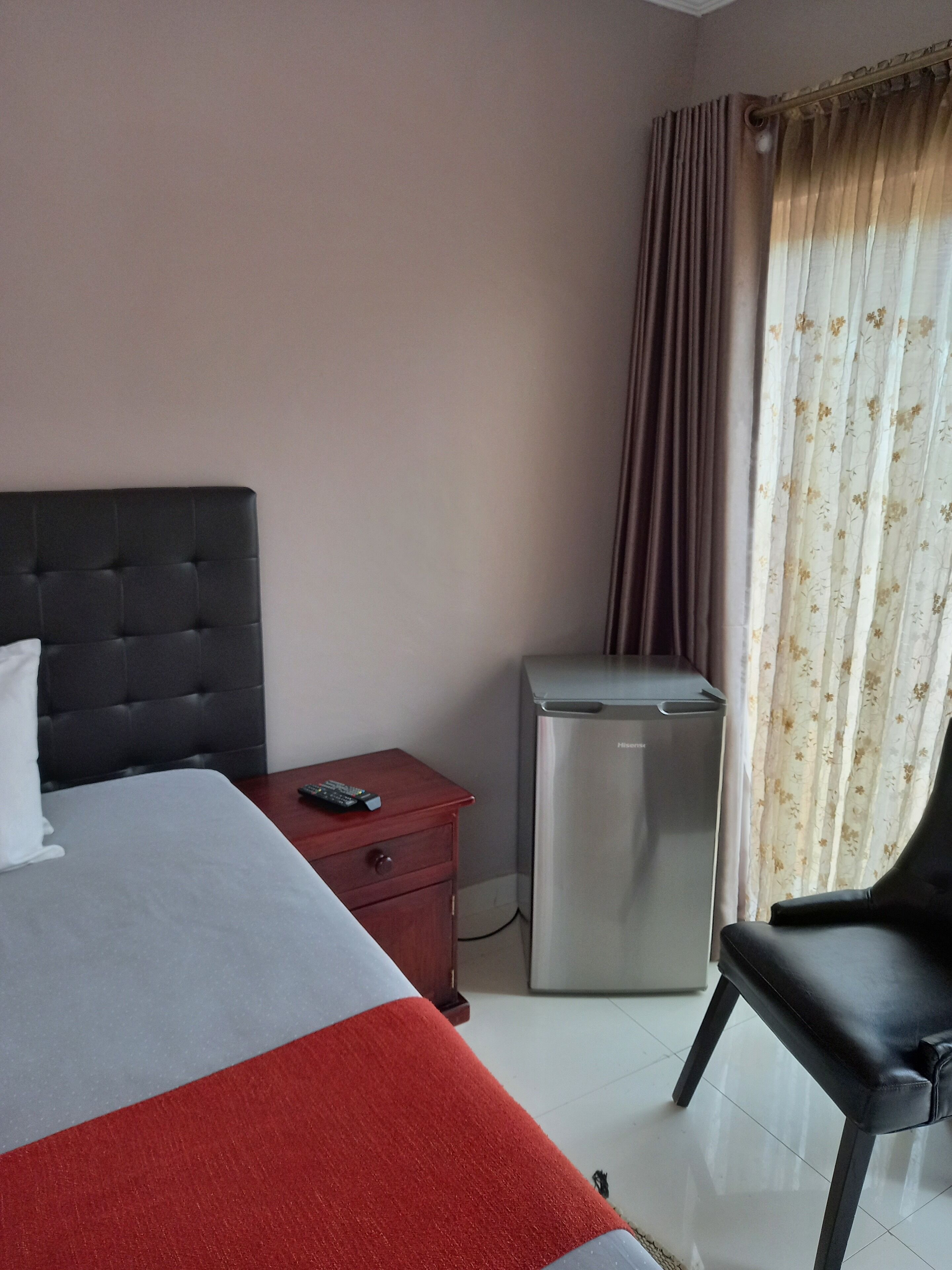A hotel room with a bed, a nightstand, a small refrigerator, a chair, and a window with curtains.