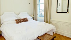 A neatly made bed with white linens and a brown patterned pillow, a wicker chair, and a large window with curtains. - Classy Historic Edinburgh Apartment (Edinburgh)