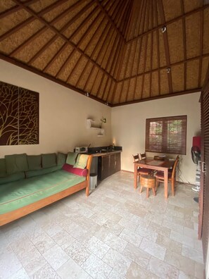A room with a thatched roof, a sofa, a dining table with chairs, a small kitchen area, and a window with blinds.