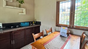A dining area with a wooden table, chairs, a window with a view, a kitchen counter, and a wall-mounted shelf with a plant. - Upper Level 1BR Apartment with Shared Pool (Ubud)
