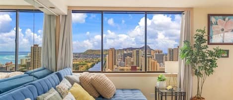 A modern living room with a large window offering a cityscape view, a blue sofa with cushions, a small table with a lamp, and a potted plant.