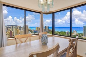 A dining area with a table and chairs, a decorative vase, and large windows offering a cityscape and ocean view.