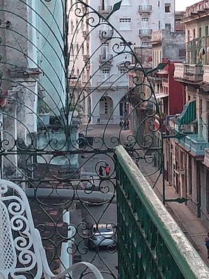 Une vue à travers une grille en métal ouvragée donnant sur une rue bordée d’immeubles, avec une voiture garée et un piéton.