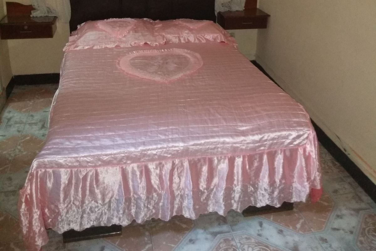 Une chambre à coucher avec un lit recouvert d’une couvre-lit rose, une tête de lit sombre et une fenêtre ornée de rideaux légers.