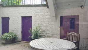 A courtyard with a round wooden table, two potted plants, and three purple doors against a white wall. - Fantastic Edinburgh Apartment (Edinburgh)