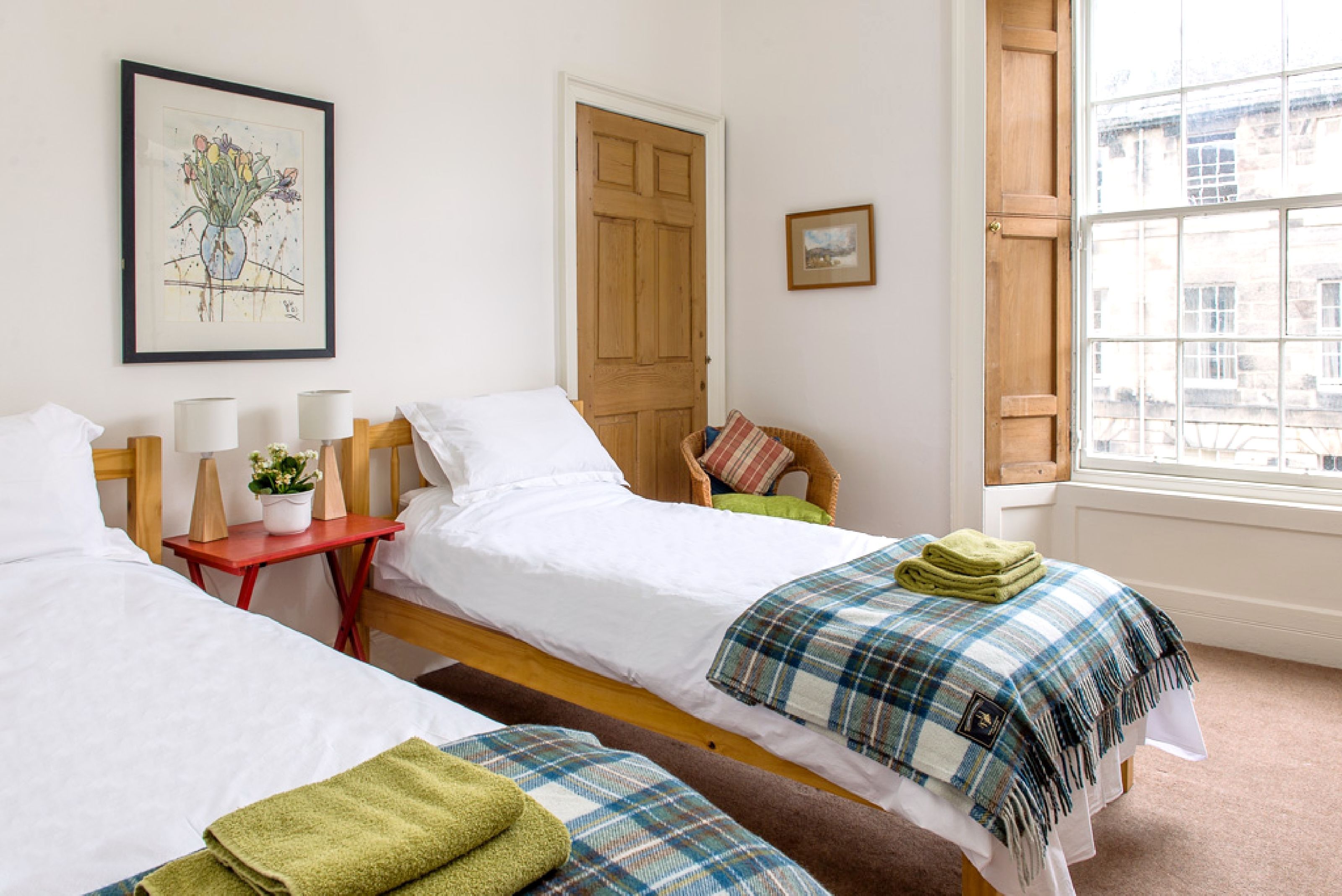 A room with two single beds, a wooden door, a window with a view, and a framed picture on the wall.