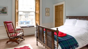 A bedroom with a large window, a wooden bed, a rocking chair, and a view of buildings outside. - Quality Stay 3-Bedroom Apartment (Edinburgh)