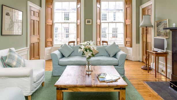 A living room with a light blue sofa, a wooden coffee table, and a vase of white flowers. - Quality Stay 3-Bedroom Apartment (Edinburgh)